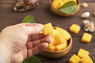 Dried and candied mango cubes in wooden bowls on brown wooden textured background with hand. Side