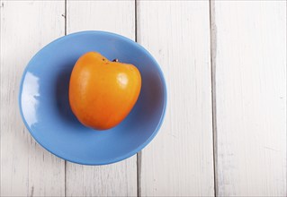 Ripe orange persimmon in a blue plate on white wooden background, with copy space