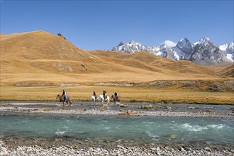 Riders riding through river in front of mountain landscape with yellow meadows, Kol Suu River and