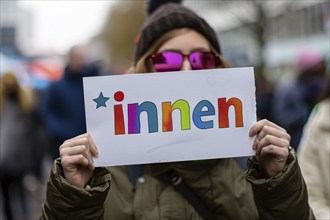 Woman holding up white sign with colorful text saying innen, a suffix used for gender neutral