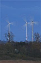 Wind energy systems turn in the evening light over a landscape framed by trees, wind turbines in