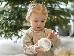 Toddler inspects a transparent Christmas tree ball in front of an illuminated Christmas tree