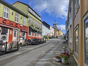 A city street with colourful houses and a church tower in the background under a partly cloudy blue