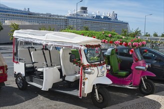 Two decorated tuk-tuks stand side by side, one white, the other pink, a large cruise ship can be