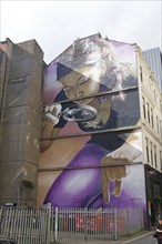 A mural shows a woman looking at something with a magnifying glass. In the background are, Mural