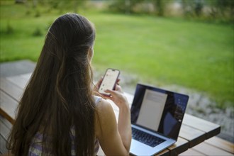 A multitasking woman using a laptop and smartphone in a serene garden setting while working on