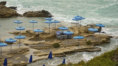 Rocks on the coast with blue parasols and deckchairs, surrounded by the sea with wavy water, Oasis