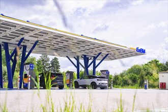 An electric car charging station under a roof with solar modules, surrounded by trees. The sky is