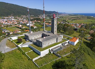 Lighthouse and other buildings in a hilly landscape with a village and the sea in the background,