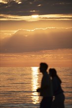 Two people walking along the sea at sunset, their silhouettes against a bright sky and calm water,