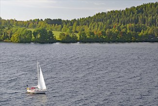 A sailboat sails on a calm lake surrounded by the green, wooded shores of Storsjon, Fröson Bridge,