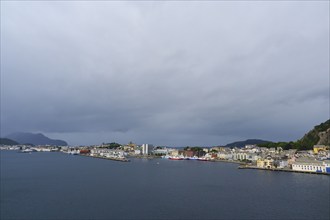 Coastal town by the sea with houses and hills in the background, boats in the harbour, cloudy sky,