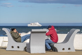 People in warm winter clothing sit on benches on the banks of the Strait of Magellan, city of Punta