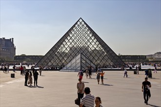 People stroll around the modern glass pyramid of the Louvre on a sunny square, Paris