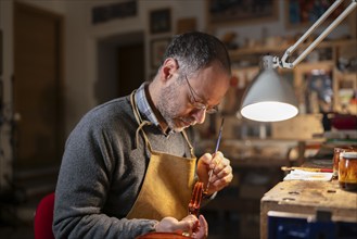 Violin maker working on a violin in his workshop, using a small brush and varnish, illuminated by a