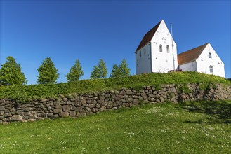 Ulbølle Kirke, whitewashed church on a hill, red roof tiles, group of trees, wall with boulders,