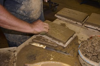 Detailed view of a worker moulding a pattern in a clay brick, surrounded by tools, Patmos Cotto