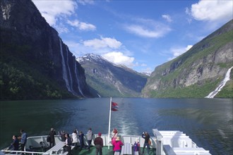 People enjoy the journey through a picturesque fjord with mountains, waterfalls and blue skies,