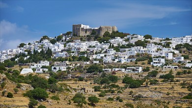 Whitewashed houses nestle against a green hill crowned by a castle under a blue sky, Monastery of