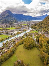 Aerial view of a village surrounded by green fields and mountains, river in the foreground, cloudy