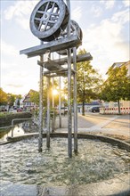 Metallic fountain in the town centre with water splashes in the evening light and autumnal trees,