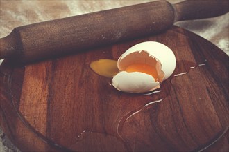 A broken egg with its yolk spilling onto a wooden cutting board sits beside a rolling pin,