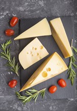 Various types of cheese with rosemary and tomatoes on black slate board on a black concrete