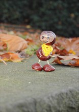 Funny chestnut figure with yellow scarf on a wall in autumn leaves, North Rhine-Westphalia,