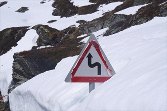 Traffic sign on a snow-covered mountain road indicating a sharp bend, June, Geiranger, Fjordland,