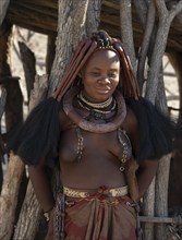 Young traditionally dressed Himba woman, leaning against a wooden hut, near Opuwo, Kaokoveld,