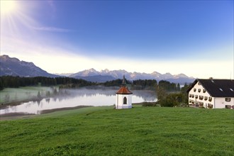 Landscape with lake, small chapel and house in the morning sunlight, framed by mountains and