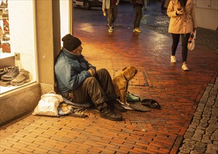 Homeless man with a dog sits in a shopping mall as people walk by in Husum, Schleswig-Holstein,