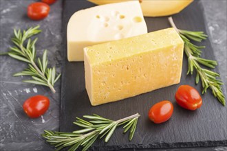 Various types of cheese with rosemary and tomatoes on black slate board on a black concrete