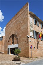 Modern museum building made of red bricks with blue shutters and the Spanish flag under a clear