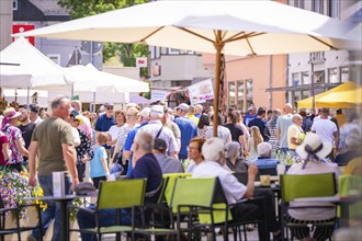 A lively market square in summer with many people sitting or walking under parasols, garden fair