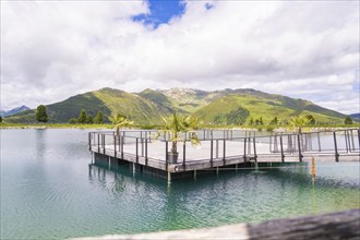 A tranquil lake with a pier, surrounded by a beautiful hilly and mountain landscape under a partly