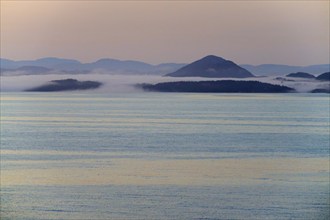 Rolling hills and islands in the morning mist at sunrise over the calm sea, Bergen, Vestland,