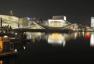 Illuminated opera house and modern buildings reflected in calm waters at night, Oslo city centre,