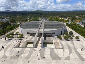 Modern architecture with a large square and people, surrounded by trees and forests, under a cloudy