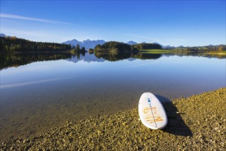 A calm lake with a surfboard on the shore, surrounded by mountains and a clear blue sky, Forggensee