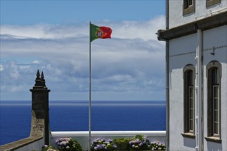 Portuguese flag flying next to a white building with a view of the blue sea and blooming flowers,