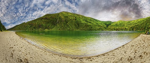 A calm lake with clear water, surrounded by green mountains and a sandy beach under a dramatic sky,