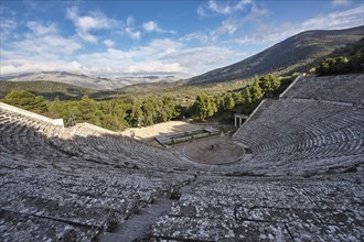 Panoramic view of an ancient amphitheatre against a mountain backdrop under a partly cloudy sky,