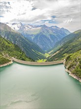 A large reservoir with a dam, surrounded by high mountains and green vegetation under a partly