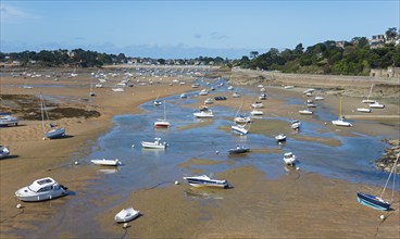 Many boats are scattered on the sandy shore at low tide, with the coast and houses behind them,