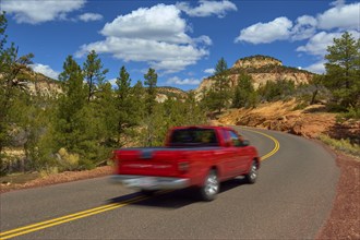 A red pickup truck drives on a winding road through a scenic mountain region with trees and blue