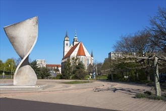 Modern sculpture in front of a church with towers and trees in the background, Monument of