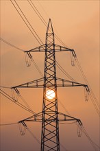 Setting sun behind silhouette of high-voltage power line pylons, shrouded in orange light amidst