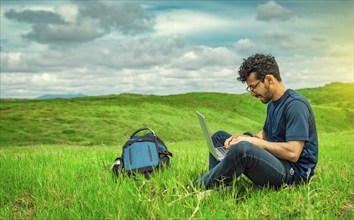 Smiling freelancer young man with laptop working from the field. Close up of man sitting on green