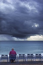 Elderly man sitting on bench at dyke and watching dark, menacing storm clouds over the sea